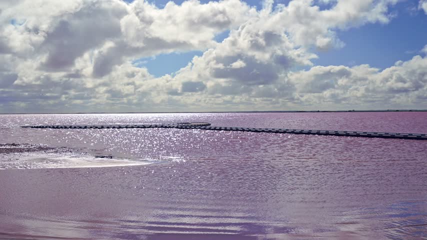 A lone woman walking along a narrow causeway dwarfed by the vast expanse of shimmering pink waters at Lake Tyrrell, beneath a blue sky adorned with white, richly textured, billowing clouds, Victoria, Australia.