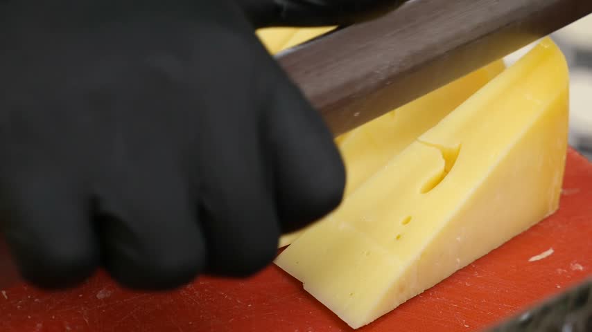 Close up video footage features person wearing black nitrile gloves carefully cutting block of yellow Swiss cheese with large knife on red plastic board for commercial food preparation.