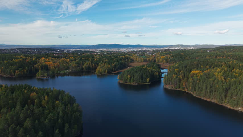 A lake bordered by mixed forest and yellowing foliage stretches toward a distant town and low hills beneath wispy clouds in Norway.