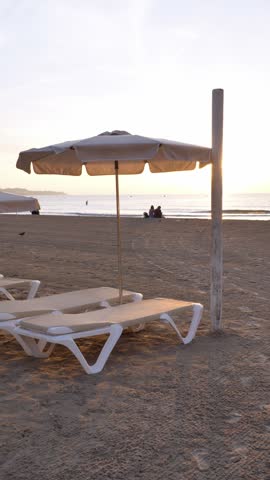 Vertical view of beach umbrellas and loungers on Postiguet Beach at sunrise, with people sitting by the shore in Alicante, Spain. Peaceful coastal morning scene.