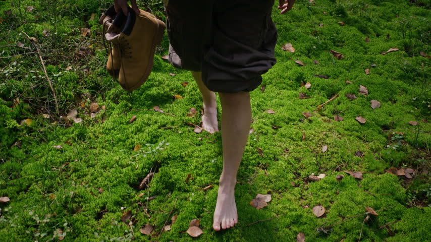 Caucasian young adult walking barefoot on green moss in forest while holding hiking boots, high angle slow motion. Footage suited for ecotherapy, grounding, and mental wellbeing campaigns