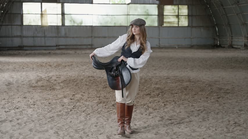 Beautiful young horsewoman carrying saddle in riding hall