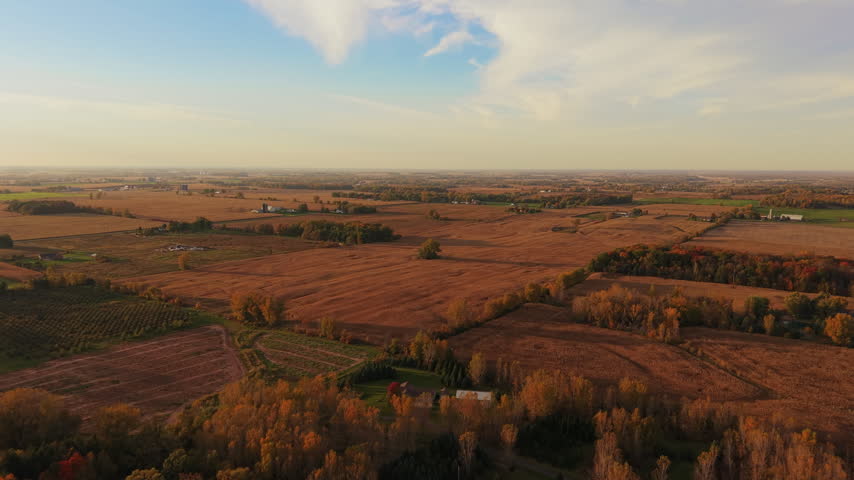 Harvested fields divided by colorful tree lines and dotted with buildings extend toward a hazy horizon beneath a partly cloudy sky in Wisconsin during the autumn