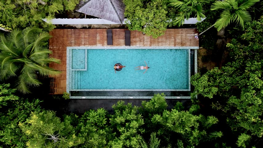 An overhead view of a couple of men and women floating in a turquoise pool surrounded by lush tropical greenery at a resort in Krabi, Thailand, during daylight. drone top view