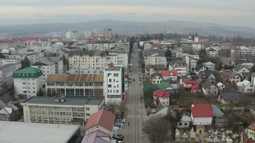 Aerial view of a town in Romania Bucovina, showing a main road flanked by residential buildings, apartment blocks, and houses with colorful roofs. The camera slowly moves forward, revealing more of