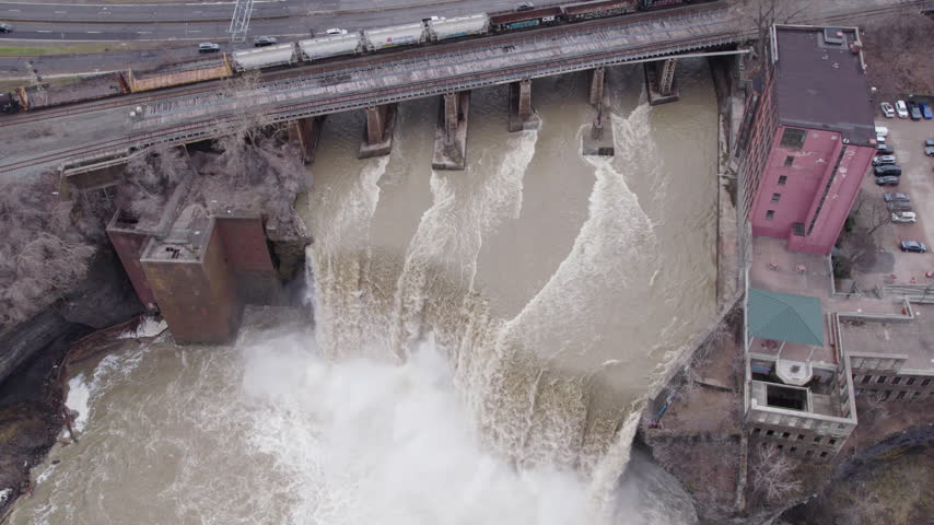 Dramatic overhead drone footage of High Falls in Rochester, New York, showing massive sheets of water plunging into the Genesee River, swirling mist, rocky cliffs, and historic industrial buildings.