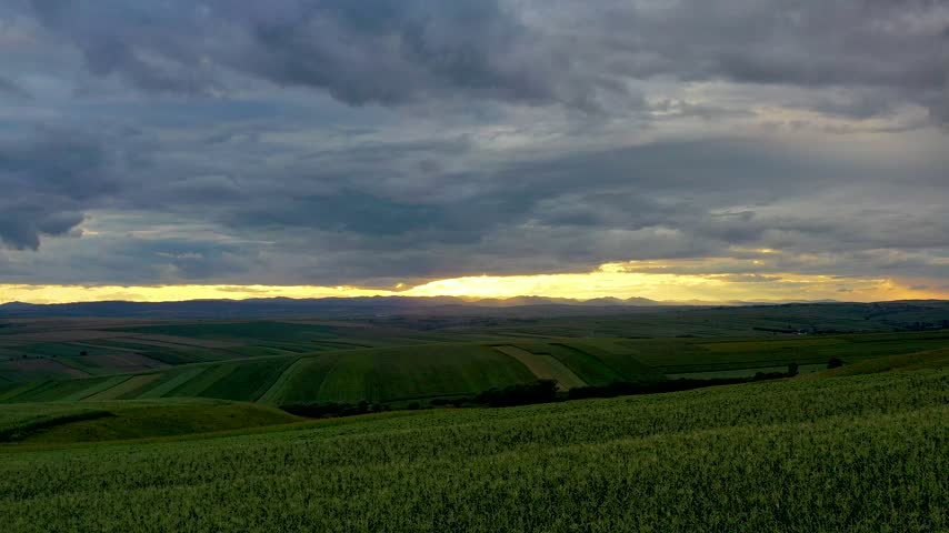 Time-lapse of a vast rolling countryside landscape in Romania Bucovina at sunset. Dark clouds dominate the sky with golden light breaking through the horizon, illuminating green fields and hills. No