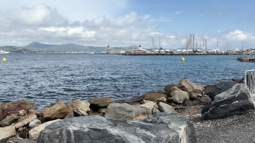 Scenic waterfront view of a Mediterranean marina in southern France, featuring luxury yachts, sailboats, a harbor pier, rocky shoreline, blue sea, and distant coastal mountains.