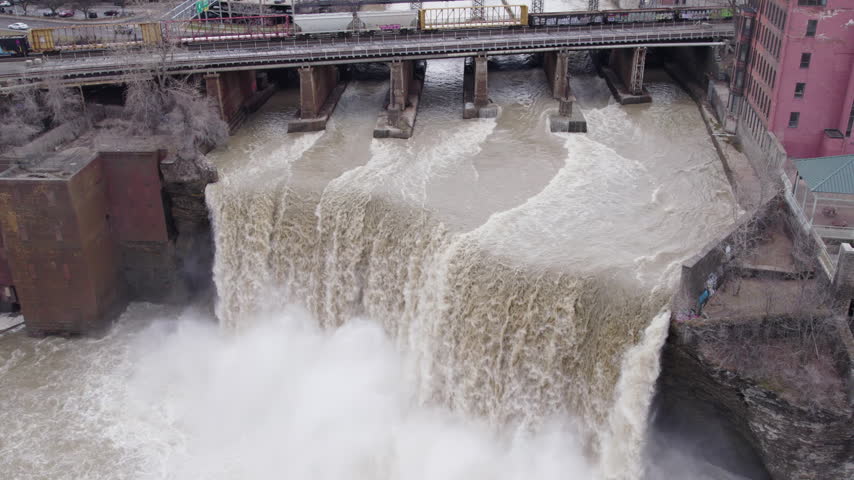 Dramatic aerial drone footage of High Falls in Rochester, New York, capturing powerful cascading water, turbulent rapids, rising mist, and a freight train crossing the railroad bridge above.