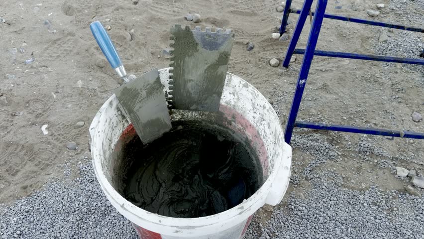 Construction tools including trowels resting in a bucket filled with wet cement on a sandy surface, with gravel and a blue structure visible in the background