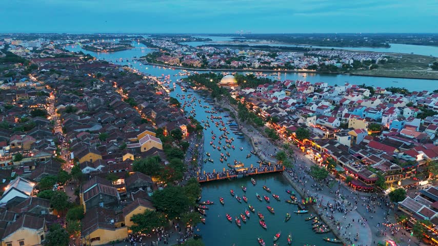 At dusk in Hoi An, Vietnam, boats glide along the river past bridges, colorful rooftops, and warmly lit streets, with the coastal city stretching to the horizon.