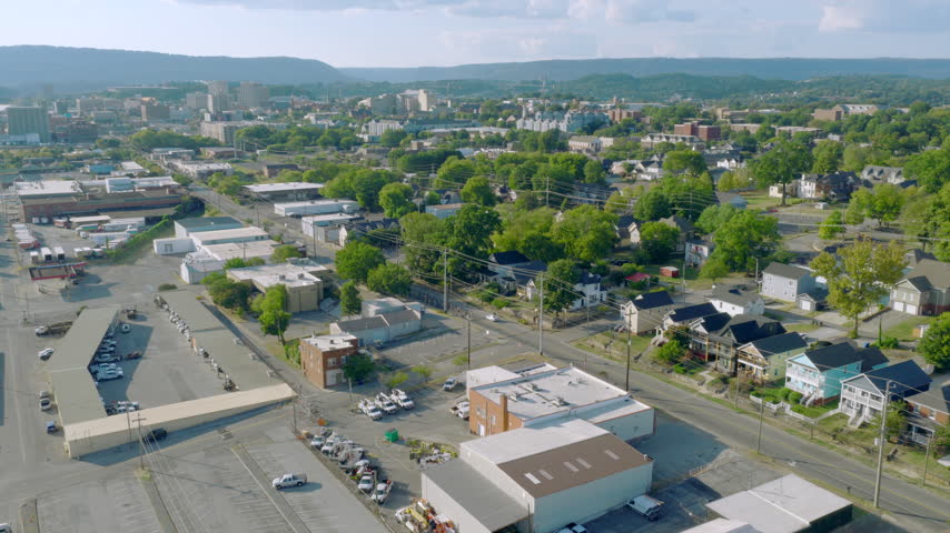 Wide aerial of a Chattanooga, Tennessee neighborhood with houses, warehouses and leafy trees leading toward downtown buildings and forested ridges on a sunny day.
