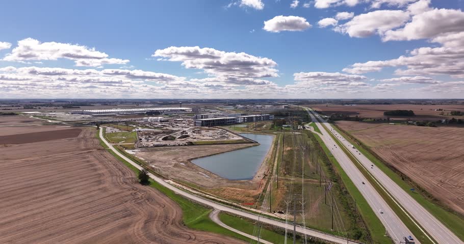 Aerial view of a large industrial construction site featuring a retention pond, adjacent to a highway, surrounded by agricultural fields in Washington Court House, Ohio, United States.