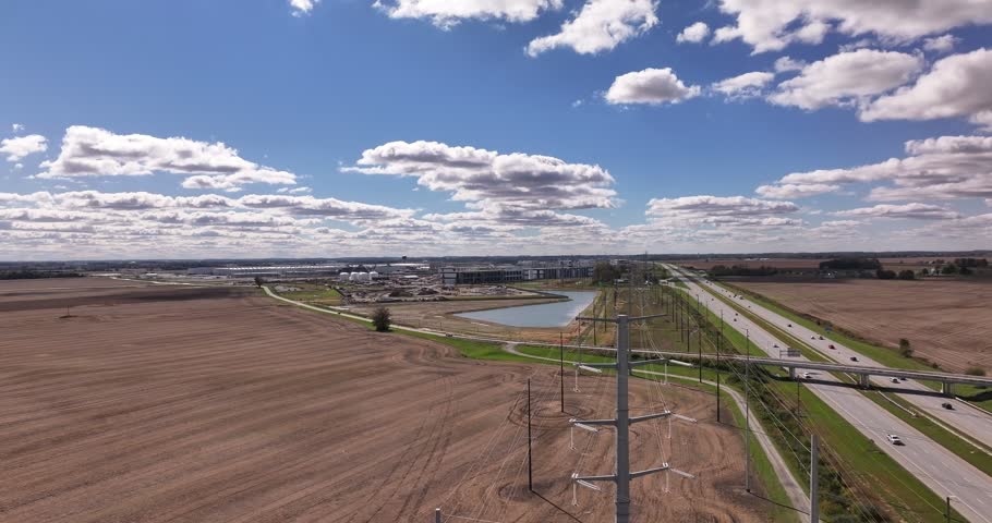 Aerial view of a highway running alongside agricultural fields, power lines, and a small pond under a blue sky with clouds in Washington Court House, Ohio, United States.