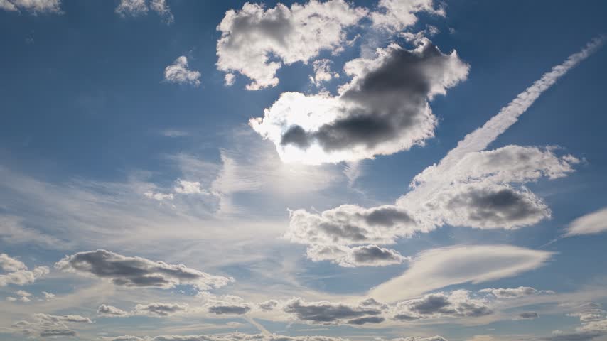 Bright sun shines behind white clouds and thin airplane vapor trails in a clear blue sky during the afternoon. Nature displays soft formations of clouds moving slowly in the atmosphere above ground.