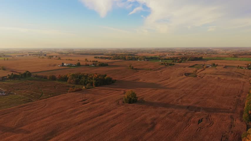 Aerial establishing shot of broad cultivated fields extending towards the horizon, divided by tree lines and grassy strips, with scattered rural buildings beneath partly cloudy sky in autumn Wisconsin