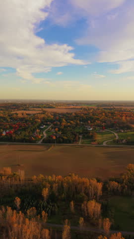 Vertical aerial shot of detached houses, lawns and curving roads sit among colorful trees beside broad agricultural fields beneath a partly cloudy sky in Wisconsin during colorful autumn season