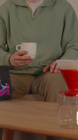 Medium shot of man hands holding coffee mug over table featuring a V60 coffee dripper and specialty coffee bag.