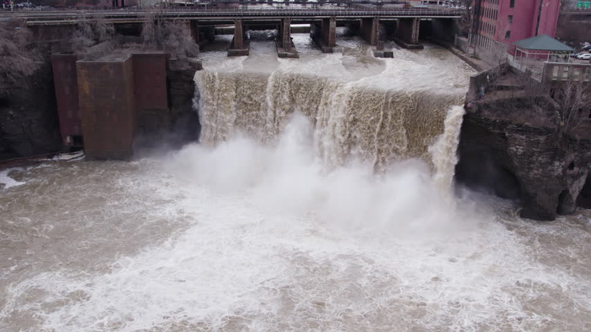 Dramatic aerial view of High Falls in Rochester, New York, showing powerful cascading water, turbulent rapids, rising mist, and historic industrial structures along the Genesee River.