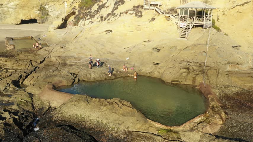 Aerial drone view of a rocky sandstone shoreline with a large tide pool and small figures exploring, late afternoon.