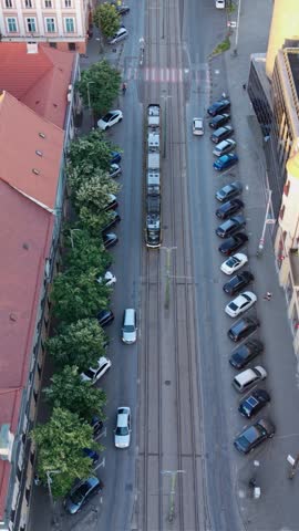 Articulated tram traveling on rails through a narrow historic street beside red tiled roofs, courtyards and parked vehicles. Vertical video of electric public transport and urban rail infrastructure.