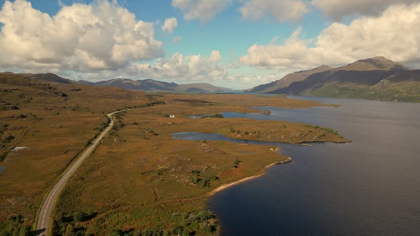 Aerial view of winding road and loch with mountains under partly cloudy sky