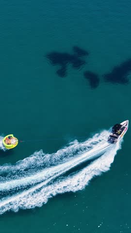 Speedboat pulling a yellow inflatable across turquoise water, with white wakes tracing a curve above dark submerged patches. Vertical video.