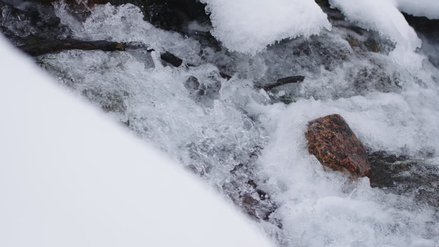 Small creek flows rapidly through snowy woodland ravine after heavy snowfall. Cold water movement prevents stream from freezing over