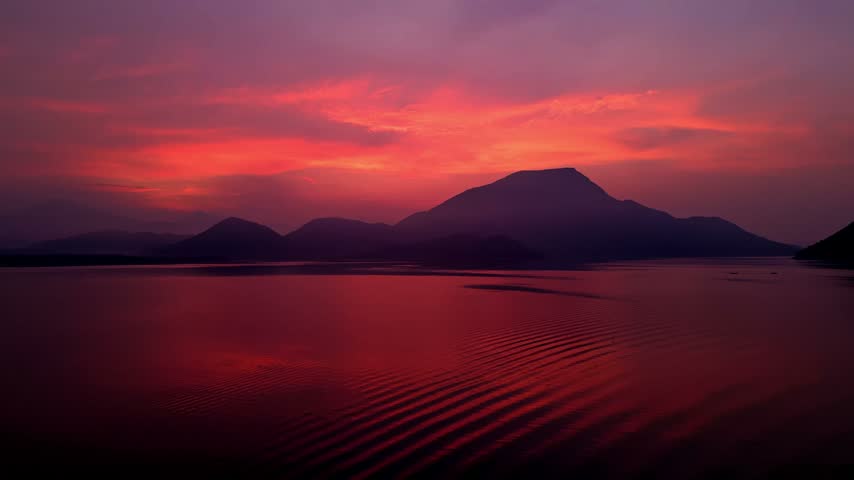 Majestic dawn sky with vibrant red and purple colors reflecting on the calm ocean water, illuminating the silhouette of distant mountains and creating a serene and picturesque natural landscape