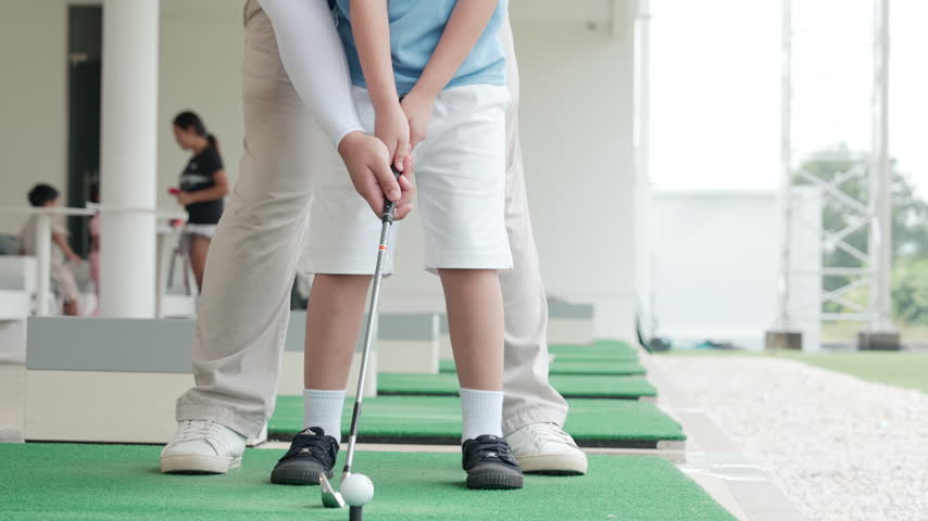 Close view of a boy holding a golf club over a ball on a practice mat as an instructor stands behind him during setup and grip training.
