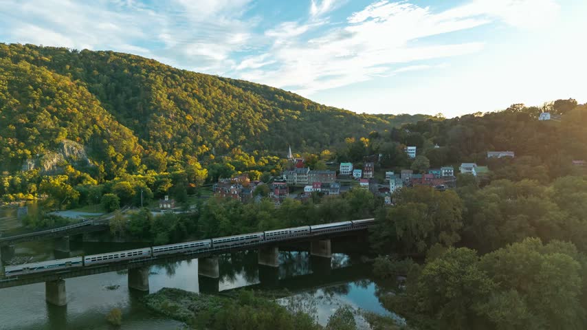 Harpers Ferry, West Virginia, aerial timelapse of a train crossing the bridge over the river past the historic town and forested mountain at sunset.