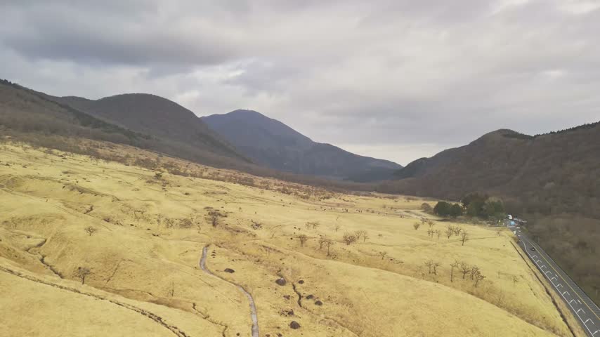 Aerial drone view of people walking along a trail in a vast mountain landscape featuring dry yellow grass and scattered trees.