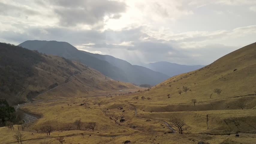 A vast aerial landscape shot capturing a dry, remote mountain valley covered in autumn grass with a road curving through it.