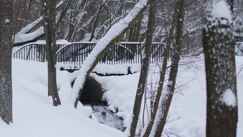 Footbridge with dark metal railings crosses snowy ravine above forest stream. Winter landscape shows woodland scene after heavy snowfall