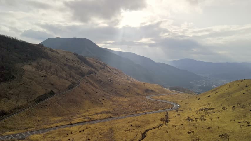 Drone shot of a winding road in a mountain valley covered in dry yellow grass under a bright cloudy sky with sunbeams.