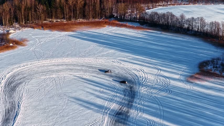 Aerial view of two cars drifting on a frozen lake, leaving tire tracks on the snow-covered surface near a forest in Latvia.