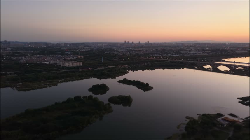 A slow aerial drift above the Luo River at sunset, with islands, mirror like water, a distant bridge and the warm afterglow along the horizon.