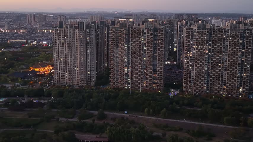 A dusk aerial drifts above a dense cluster of residential towers in Luoyang, with a green park, roads and the last light on the horizon.