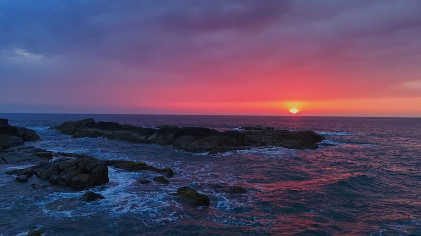 Sri Lanka - 05 05 2024: Vivid red sunset over a calm ocean horizon, with glowing reflections on the water and a tropical beach foreground featuring traditional fishing boats and rocks.
