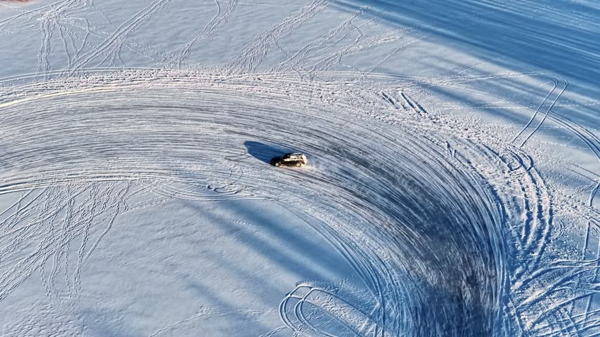 Aerial view of a car drifting on a vast snow-covered field, creating deep tire tracks in the white landscape in Latvia.