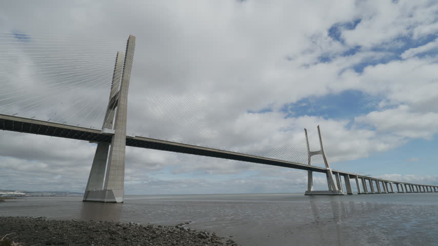 Vasco Da Gama Bridge Rises Over Lisbon Waters, Modern Cable Stayed Bridge With Long Roadway And Tall Pylons. Cloudy Sky Frames Wide River View.