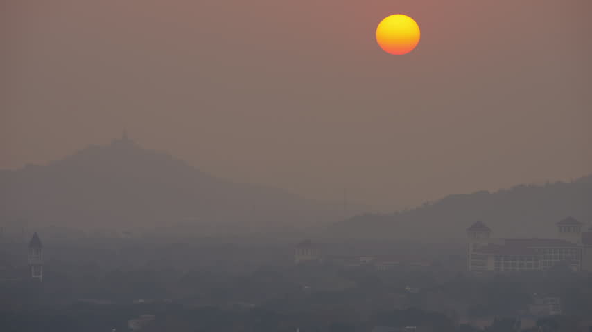 An orange red sun setting through thick haze over distant hills and rooftops
