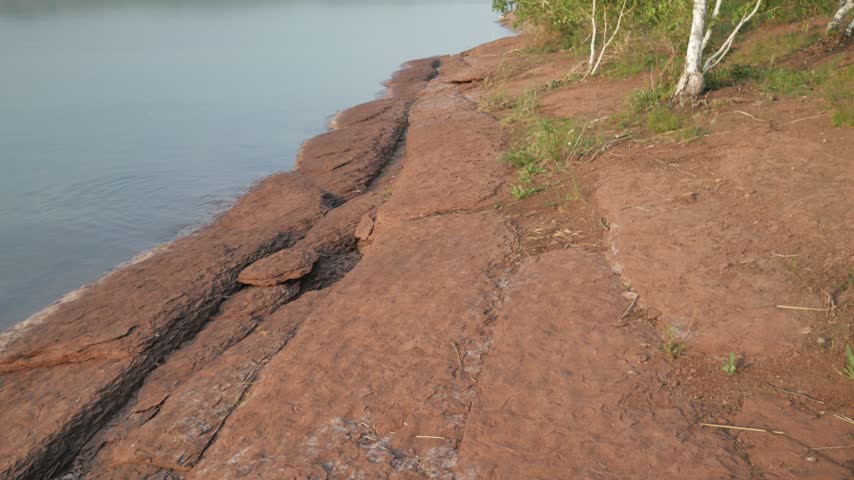 Terraced red sandstone formations line the peaceful shore of Lake Shira beneath a grassy bank dotted with birch trees. Camera glides along the stony lakeside.