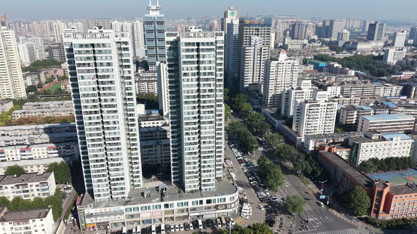 The camera circles two tall white apartment towers, revealing a school sports field, busy intersections, tree-lined avenues and a dense city skyline in clear autumn daylight.