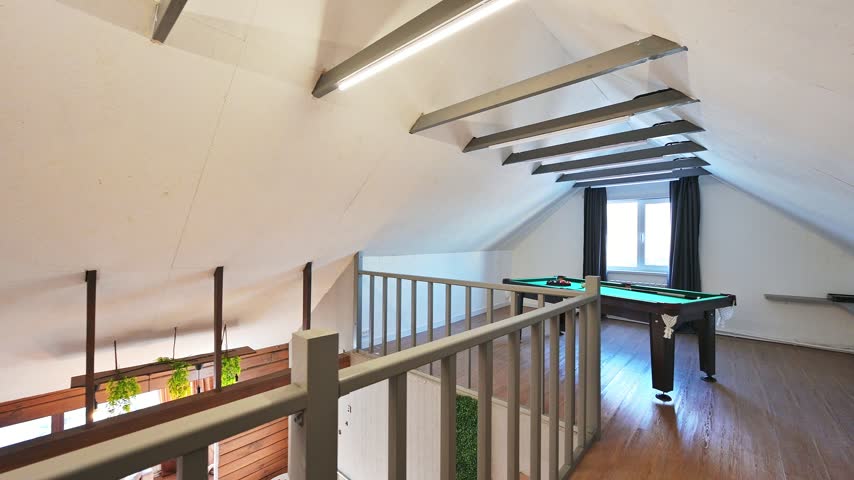 Attic billiard room with slanted ceiling, green table, wooden railing, exposed beams, window with dark curtains, minimalist finish.
