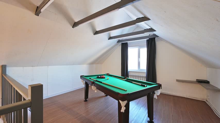Attic billiard room with green table, cues, balls, slanted ceiling, exposed beams, window with dark curtains, radiator, minimalist finish.
