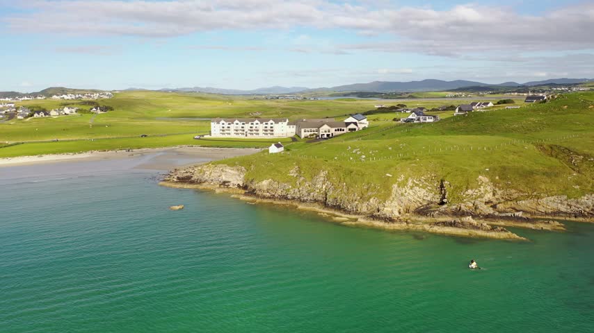 Aerial view of a hotel on the grassy headland overlooking the sandy beach and turquoise waters of the Atlantic Ocean in Downings, County Donegal, Ireland.