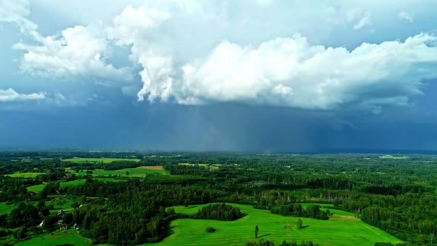 Aerial view of a massive storm cloud hovering over lush green fields and forests in Latvia.