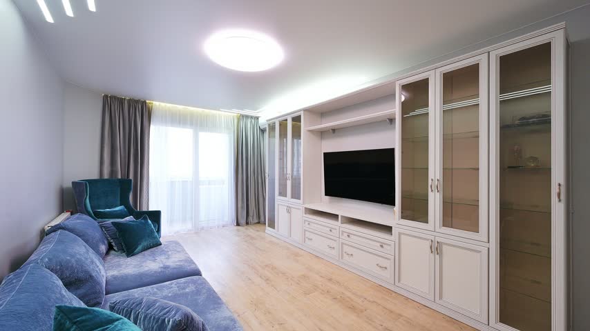 Stylish living room with blue velvet sofa, teal armchair, white built‑in cabinet, glass doors, TV, sheer curtains, soft lighting.
