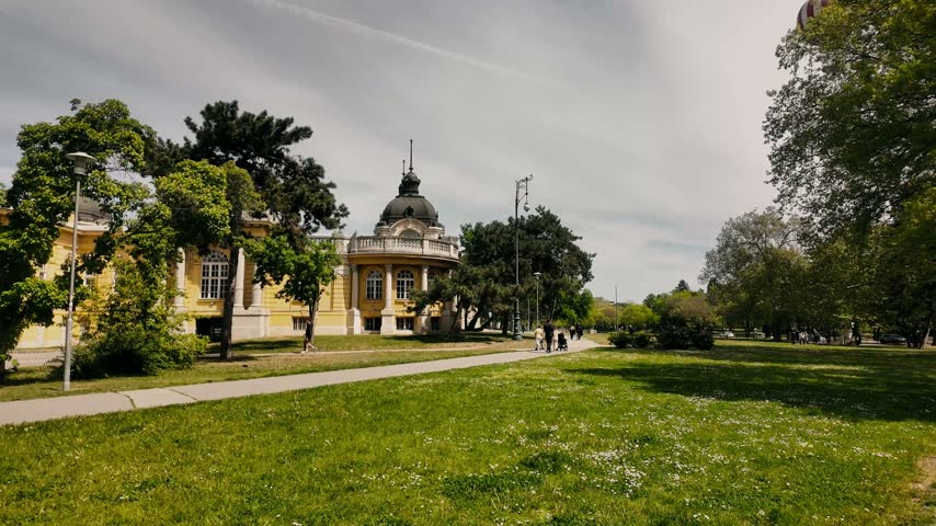 Historic yellow palace building in Budapest city park with green lawn and walking path, spring day with people strolling — European travel and architecture footage